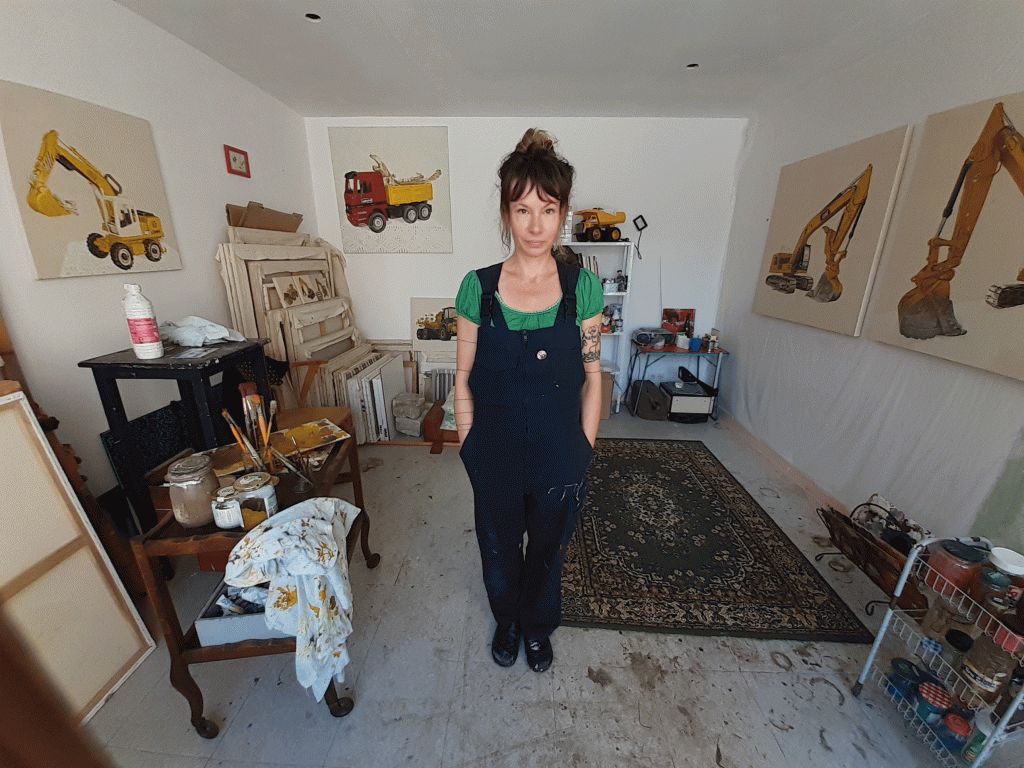 Photographie de l'artiste peintre Peggy Fraquet debout face camera au milieu de son atelier avec aux murs quelques unes de ses oeuvres.